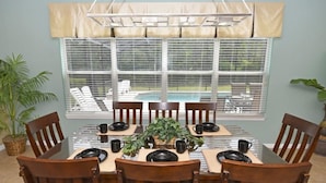 A dining area with a table set for six, surrounded by wooden chairs, a hanging light fixture, and a view of a pool through large windows. - 112 Watersong House 6 Bedroom by Florida Star (Davenport)