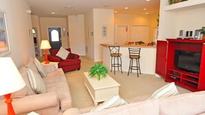 A living room with a sofa, a coffee table, a TV stand, and a kitchen area in the background.