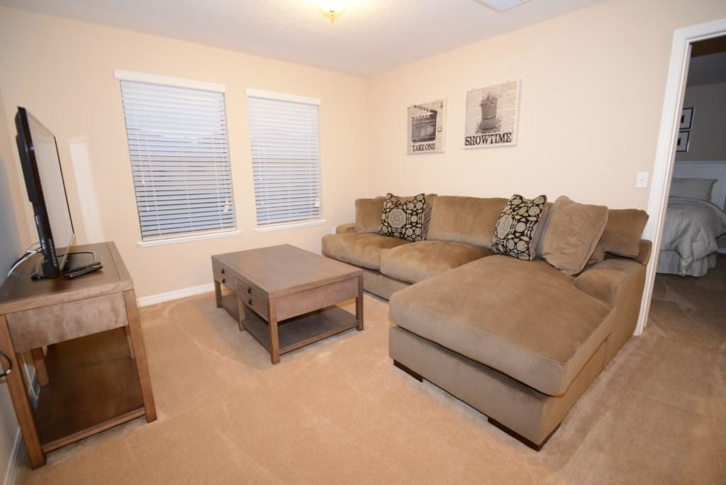 A living room with a beige sofa, two side tables, and a coffee table. There are two windows with blinds, a ceiling light, and two wall-mounted pictures.