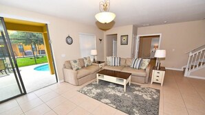 A living room with a beige sofa, a coffee table, and a patterned rug. There is a staircase to the right and a door leading outside.