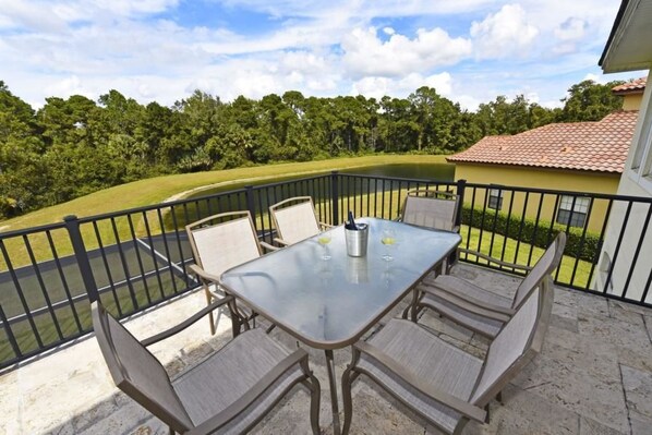 Un balcon avec une table et des chaises en verre, donnant sur un lac et une végétation luxuriante.