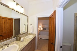 Une salle de bain dotée d’un grand miroir, de deux lavabos et d’une porte en bois donnant sur une chambre.