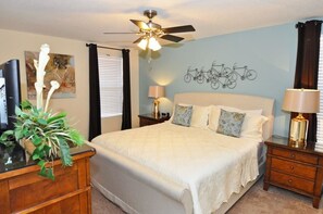 A bedroom with a bed, a ceiling fan, a wall-mounted bicycle art piece, a bedside table with a lamp, and a plant on a dresser.