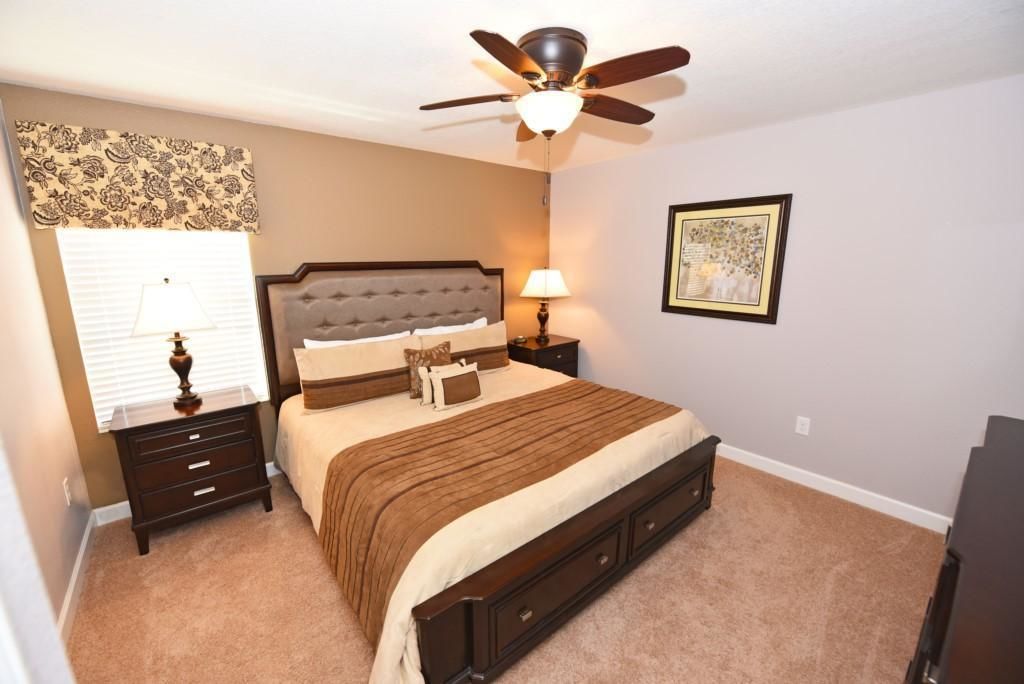 Une chambre à coucher comprenant un lit, des tables de chevet, un ventilateur de plafond, une fenêtre avec des stores et un tableau encadré accroché au mur.