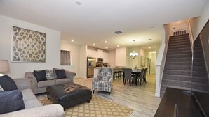 A modern living room with a sofa, ottoman, and patterned rug. A staircase leads to a dining area with a table and chairs. - 1600 Champions House 5 Bedroom by Florida Star (Davenport)