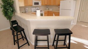 A kitchen with wooden cabinets, a white refrigerator, a microwave, and a sink. There are two black stools and a potted plant. - 16708 High Grove House 4 Bedroom by Florida Star (Clermont)