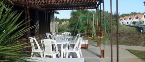A covered outdoor seating area with tables and chairs, a swing set, and a view of houses in the background.