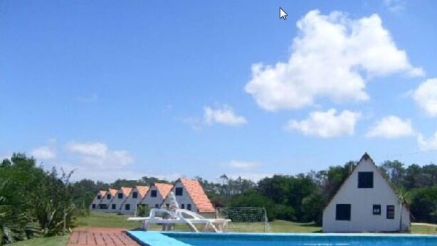 Un área de piscina con cubierta de madera, un edificio blanco con tejado rojo y un cielo azul despejado.