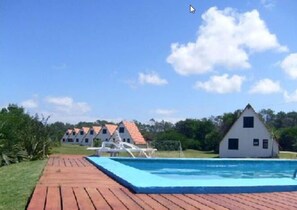 A pool area with a wooden deck, a white building with a red roof, and a clear blue sky. - Complejo Turistico Anaconda (La Paloma)