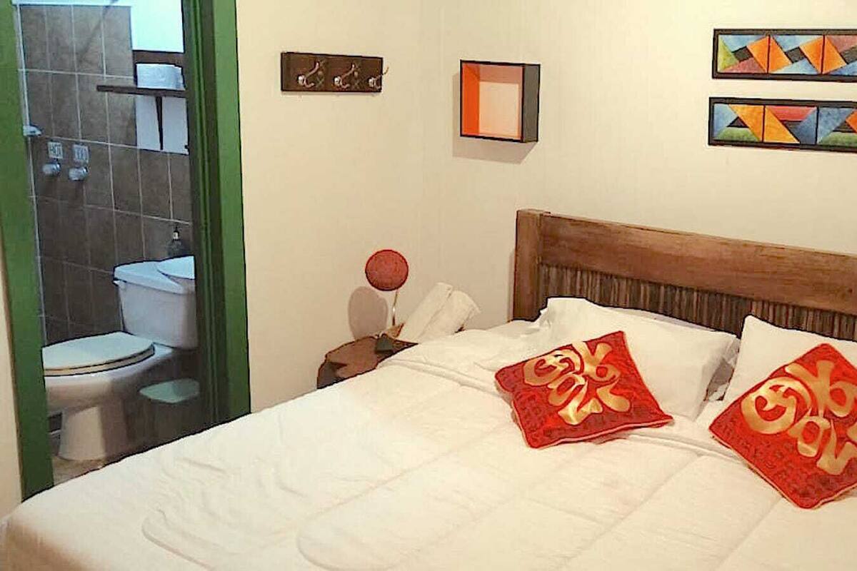 Une chambre d’hôtel avec un lit, deux oreillers aux motifs rouges et dorés, une tête de lit en bois et un mur orné de deux œuvres d’art encadrées.