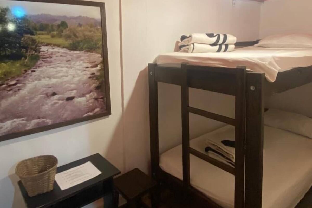 Un lit superposé avec un matelas blanc et une tête de lit, une petite table en bois avec un panier, et une image encadrée représentant une rivière accrochée au mur.