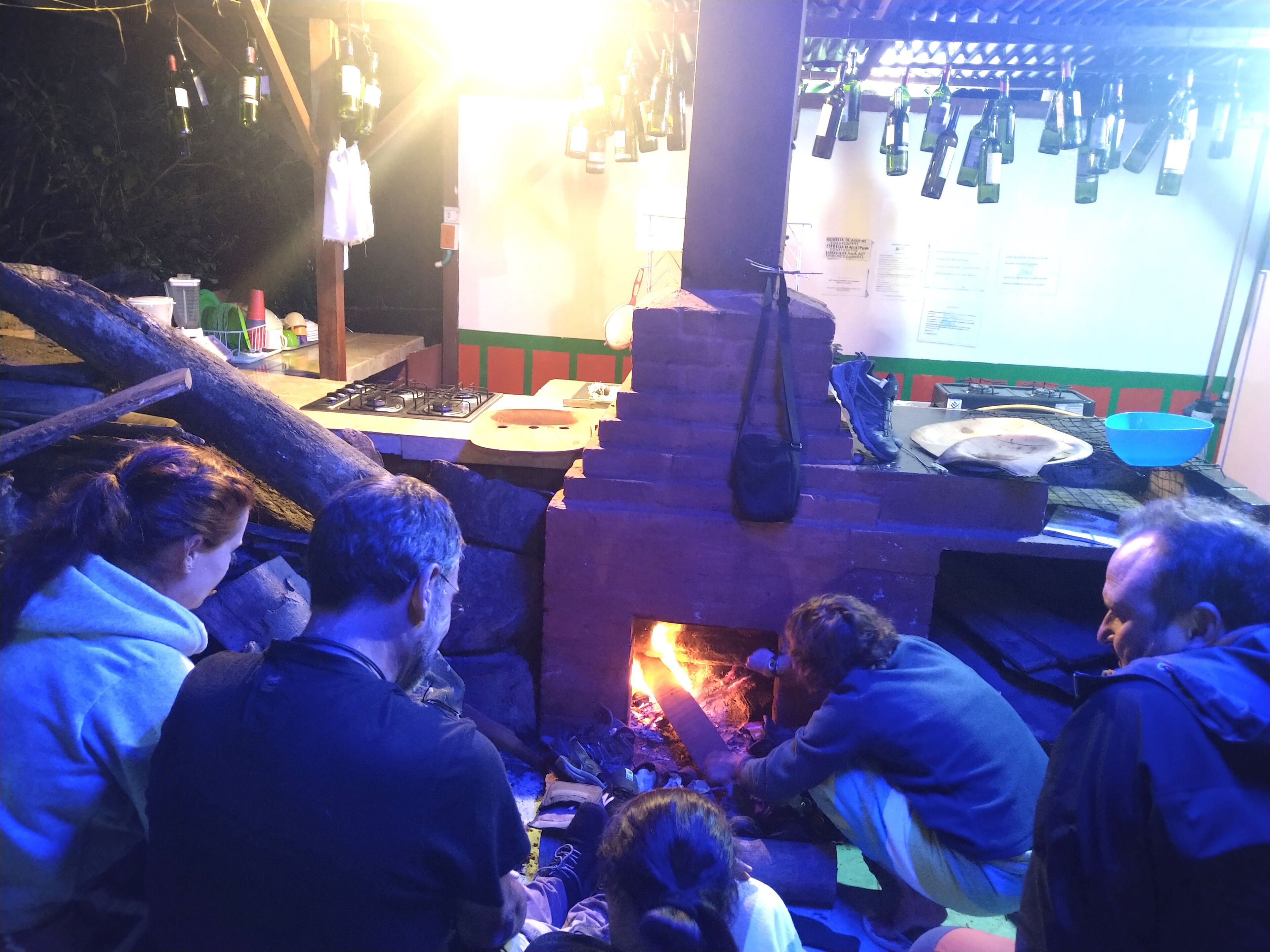 A group of people gathered around a fire pit in a rustic setting with a brick oven and hanging wine bottles.