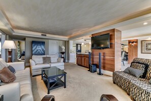 A spacious living area with a sofa, a coffee table, and a patterned ottoman. A flat-screen TV is mounted on the wall, and there is a kitchen area visible in the background.
