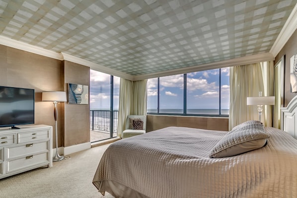 A hotel room with a large bed, a flat-screen TV, a desk with drawers, a lamp, and a view of the ocean through large windows.