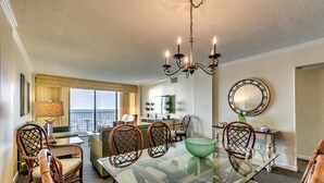 A dining area with a glass table, wicker chairs, a chandelier, and a view of the outdoors. - Carolina Dunes 304 2 Bedroom Condo by Luxury (Myrtle Beach)
