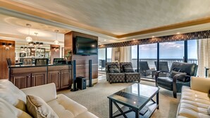 A spacious living room with a large sofa, a coffee table, and a TV mounted on the wall. There is a balcony with a view of the ocean. - Carolina Dunes 304 2 Bedroom Condo by Luxury (Myrtle Beach)