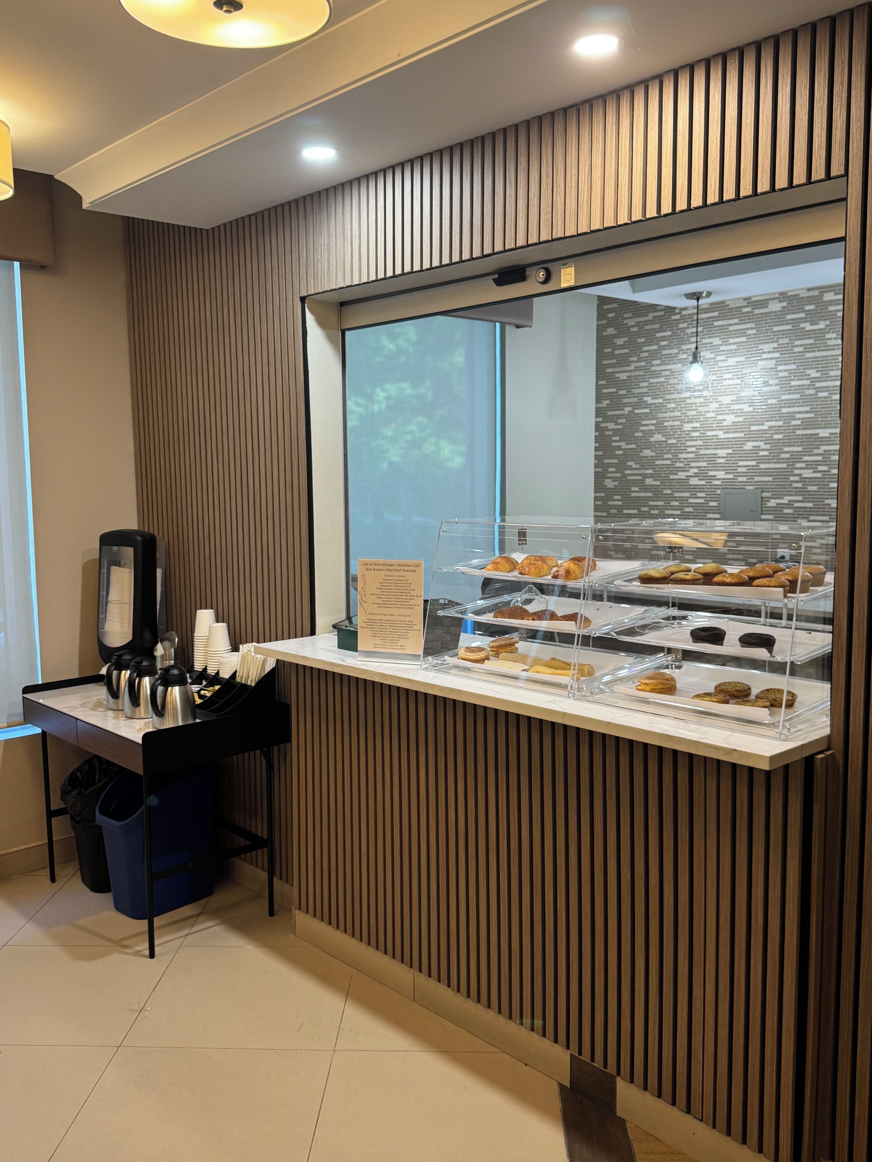 A bakery display case with various baked goods, a coffee station with cups and a coffee maker, and a wooden panel wall.