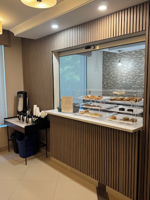 A bakery display case with various baked goods, a coffee station with cups and a coffee maker, and a wooden panel wall. - Best Western Plus Hotel Montreal (Montreal)