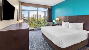 A hotel room with a large bed, a desk, a flat-screen TV, and a view of the cityscape through large windows.