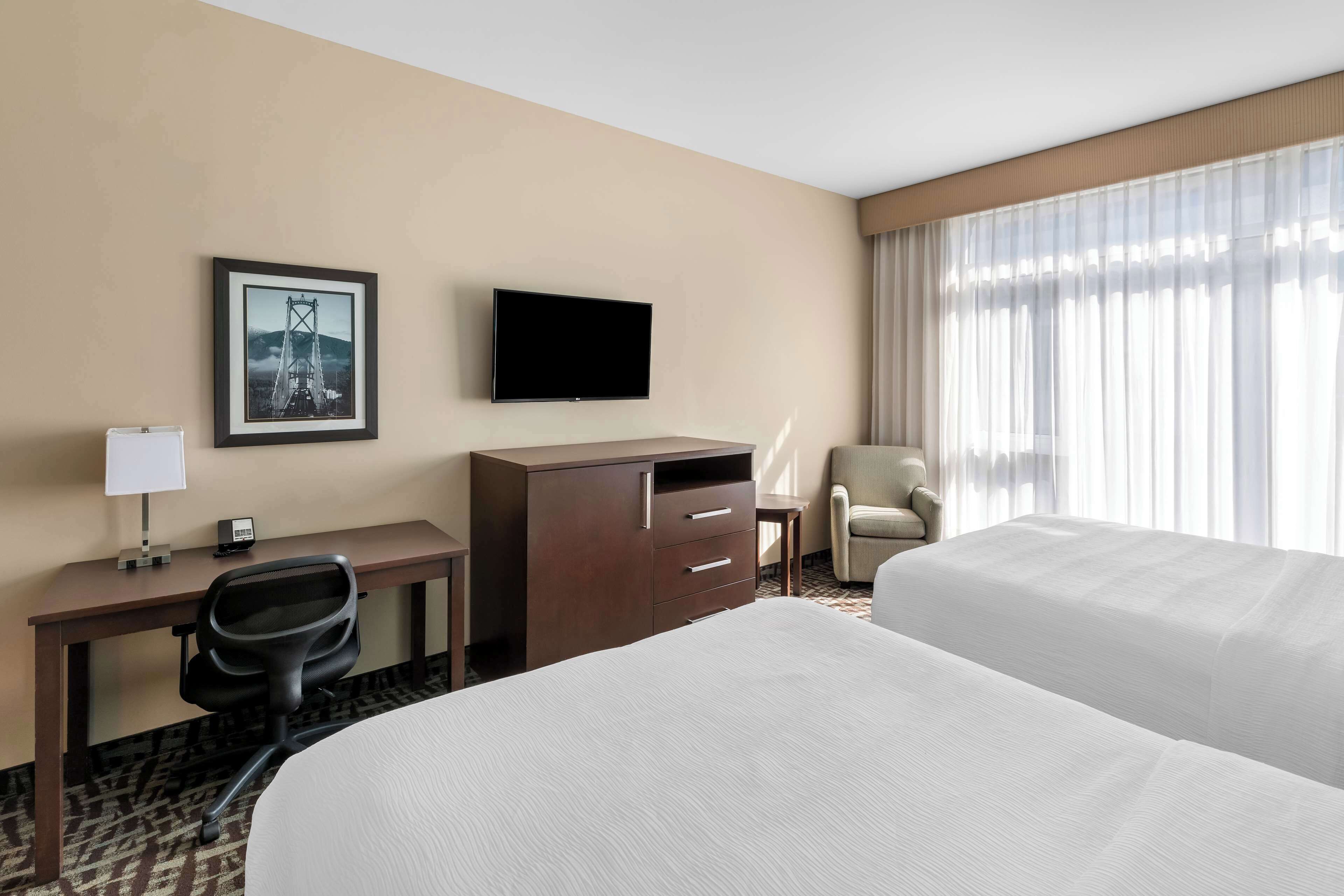 Habitación de hotel con cama, escritorio y silla, televisión, un cuadro en la pared y ventana con cortinas.
