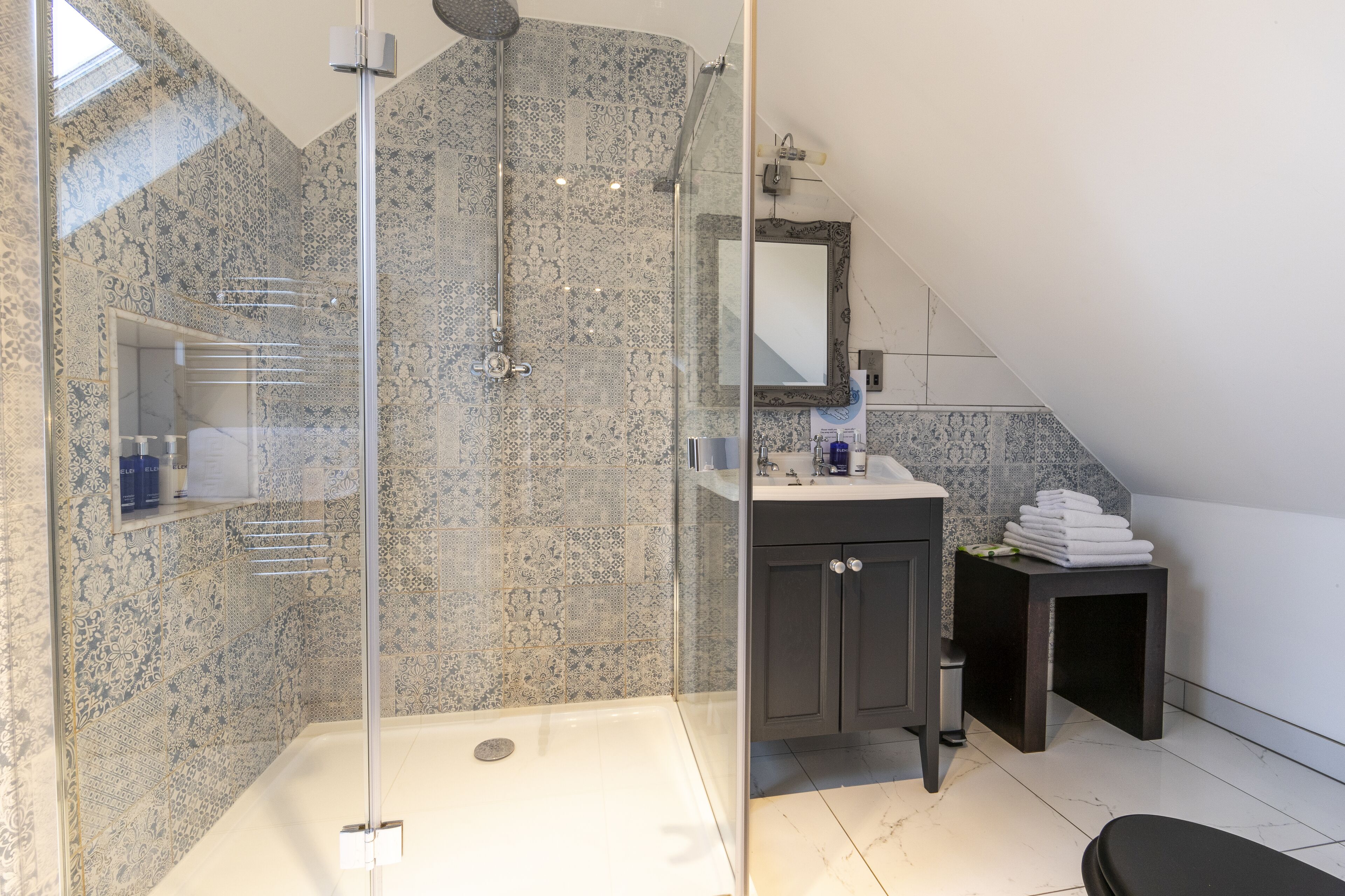 A modern bathroom with a glass-enclosed shower, a vanity with a mirror, and a small stool with towels.