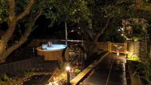 Un jacuzzi bajo un árbol, rodeado por una terraza de madera y un sendero de piedra.