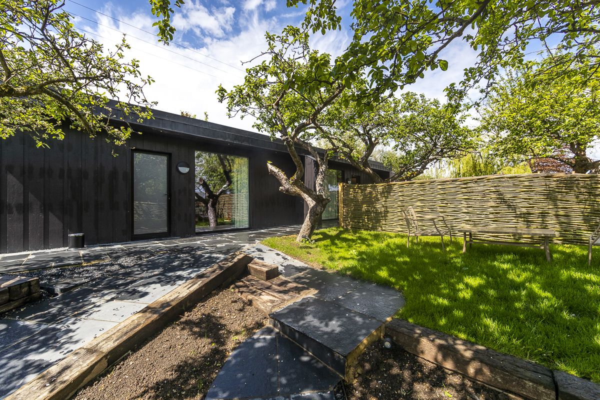 Un bâtiment moderne à la façade sombre, avec une allée pavée et un espace verdoyant agrémenté d’un arbre et d’une clôture en bois.