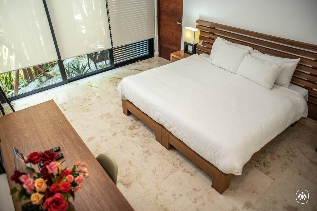A bedroom with a large bed, a wooden nightstand with a lamp, a desk with a vase of flowers, and a view of the outdoors through a sliding door.