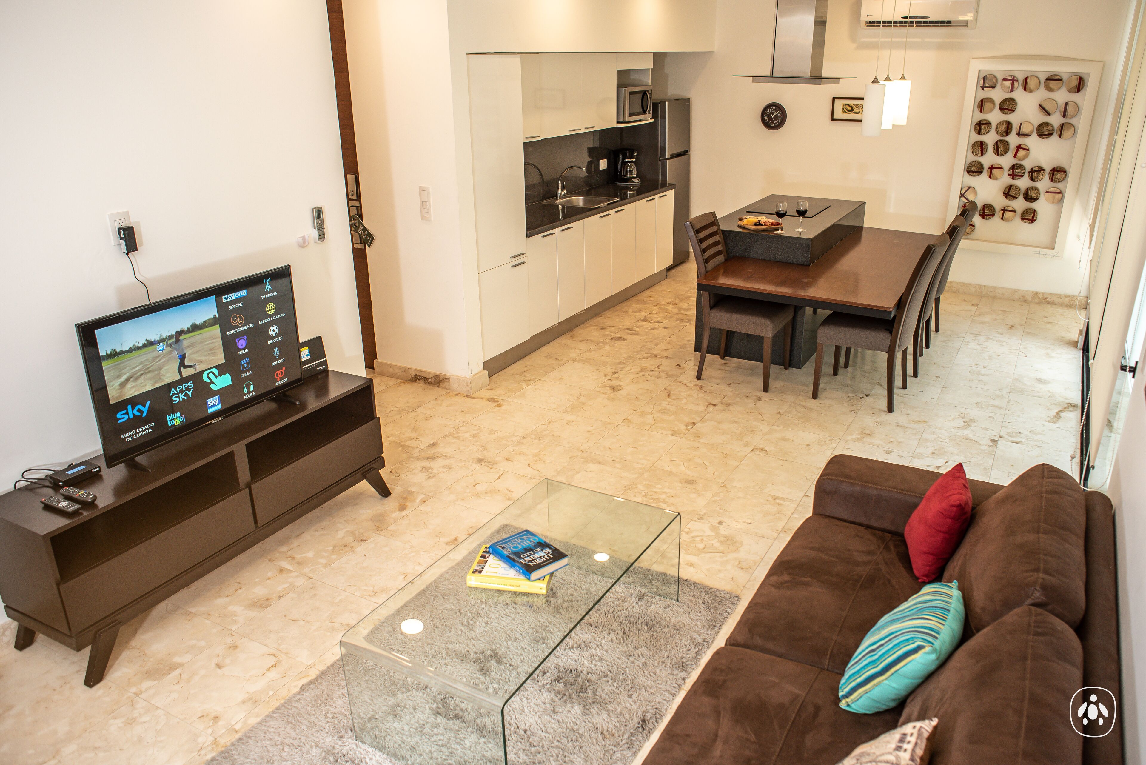 Una sala de estar moderna con un televisor de pantalla plana, una mesa de centro de vidrio, un sofá marrón con cojines, un comedor con una mesa y sillas, y una cocina al fondo.