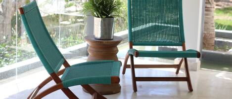 Two green wicker chairs with wooden frames, a potted plant on a wooden table, and a view of greenery through a glass door.