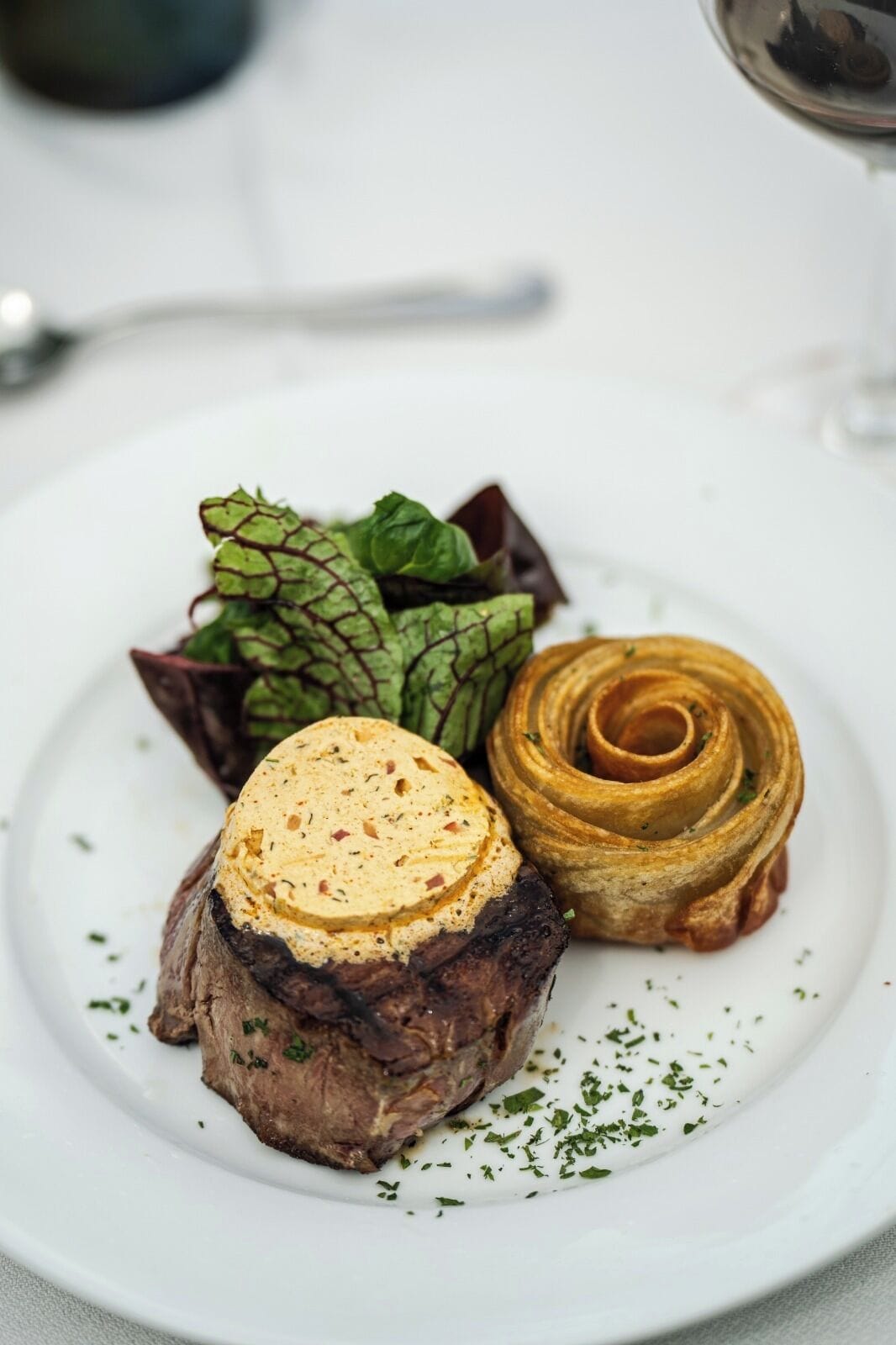 A plate with a steak topped with a creamy sauce, a spiral pastry, and a side of greens.