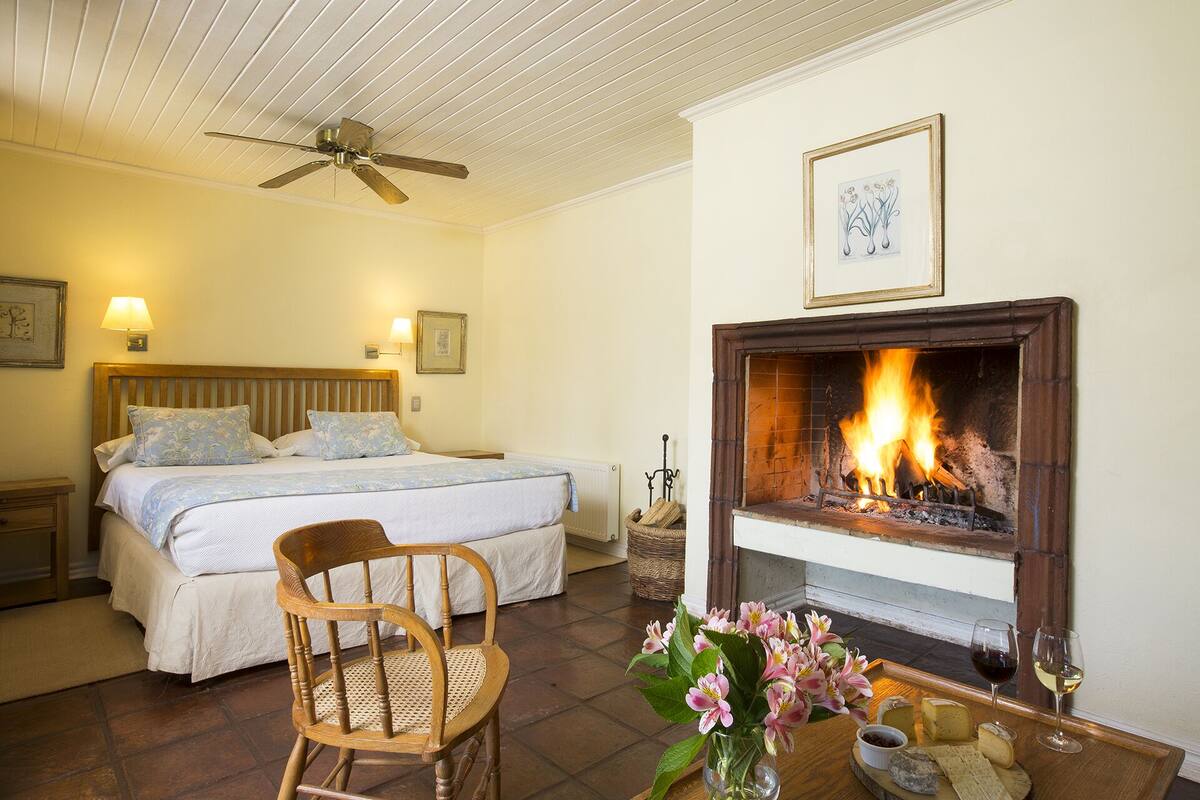 Un dormitorio con una cama, una silla de madera, una chimenea encendida y una mesa con flores y queso.