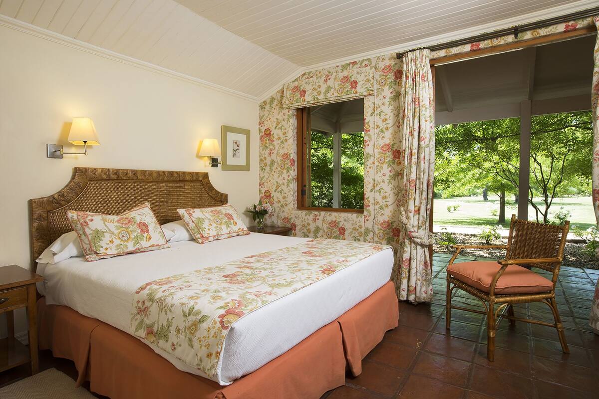Un dormitorio con una cama grande, dos lámparas de noche, un ventanal con cortinas florales y una silla de mimbre junto a la ventana.