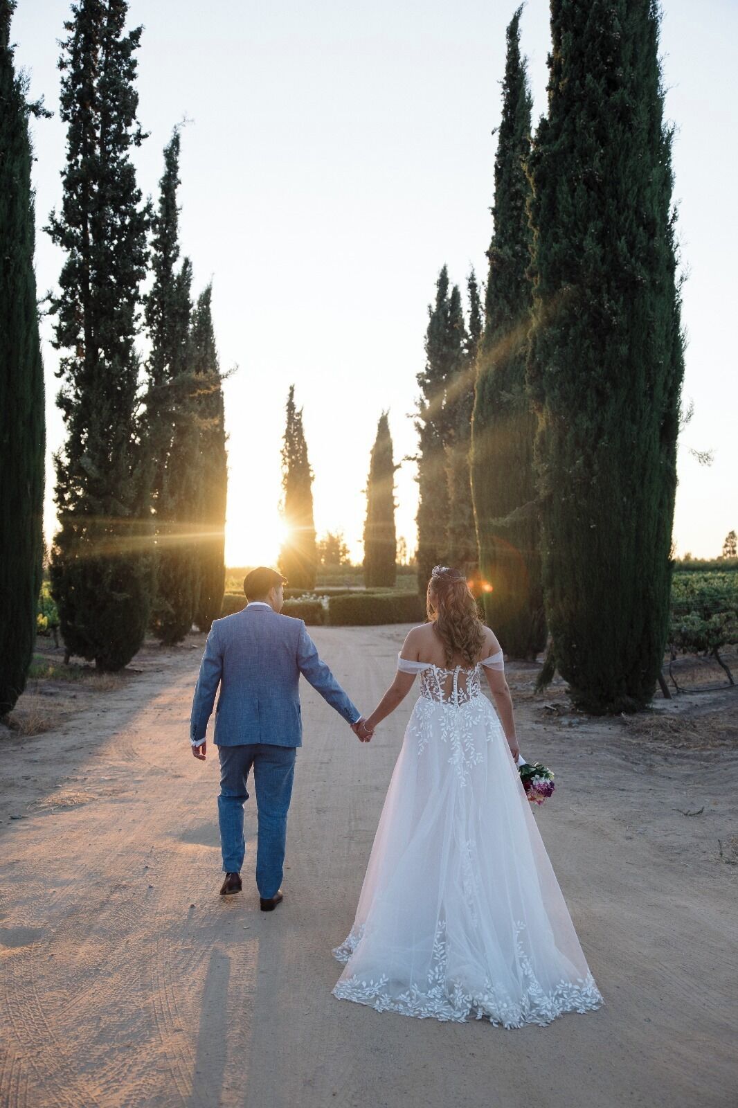 Una pareja caminando por un sendero de tierra rodeado de árboles altos, con el sol poniéndose al fondo.