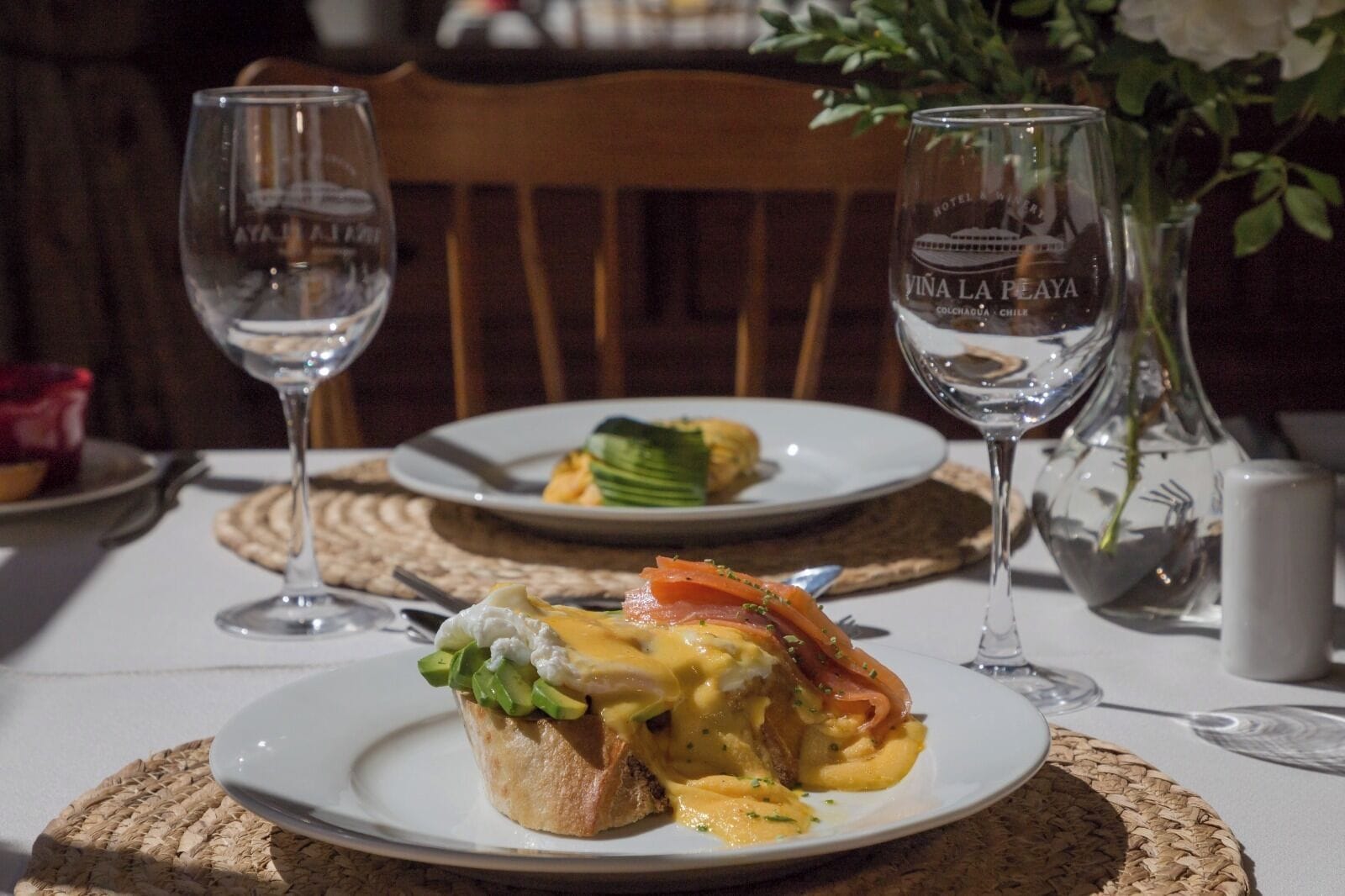 A dining table with plates of food, two wine glasses, and a vase with flowers.
