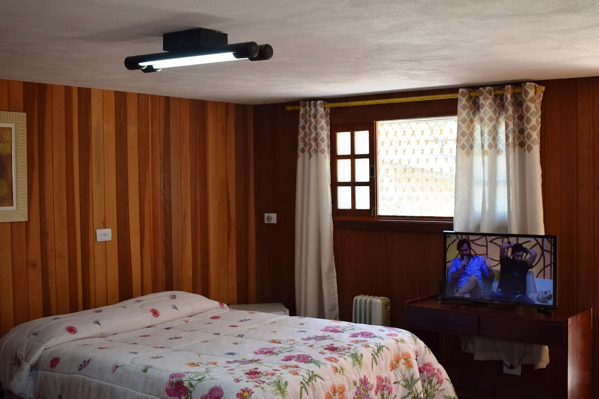 Ein Zimmer mit Holzwänden, einem Bett mit Blumenmuster auf der Bettwäsche, einem Fernseher und einem Fenster mit gemusterten Vorhängen.