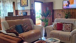 Un salón con un sillón de cuero marrón, un sofá color crema y una mesa de centro redonda. Hay un televisor montado en la pared y una puerta que da a un área exterior.