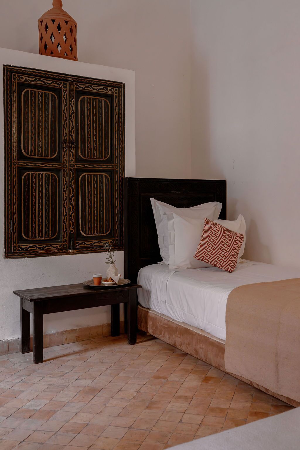 A bedroom with a bed, pillows, a nightstand, a decorative wall piece, and a tiled floor.