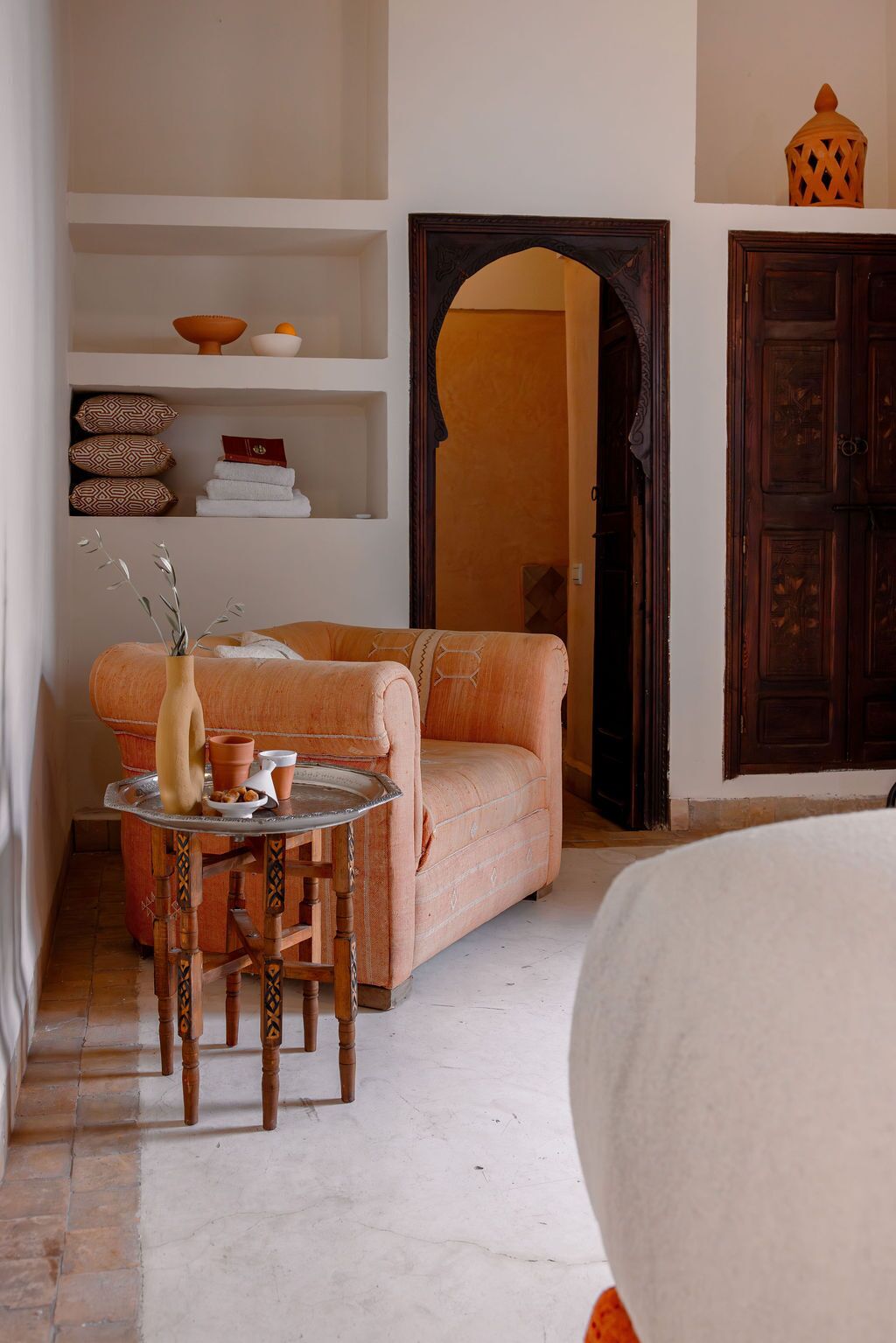 A room with a sofa, a small table with a vase and a pitcher, and a shelf with patterned pillows and a book.