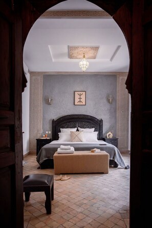 A bedroom with a large bed, a bench, a nightstand, and a wall-mounted light fixture. - Les Sources Berbères Riad & Spa (Marrakech)