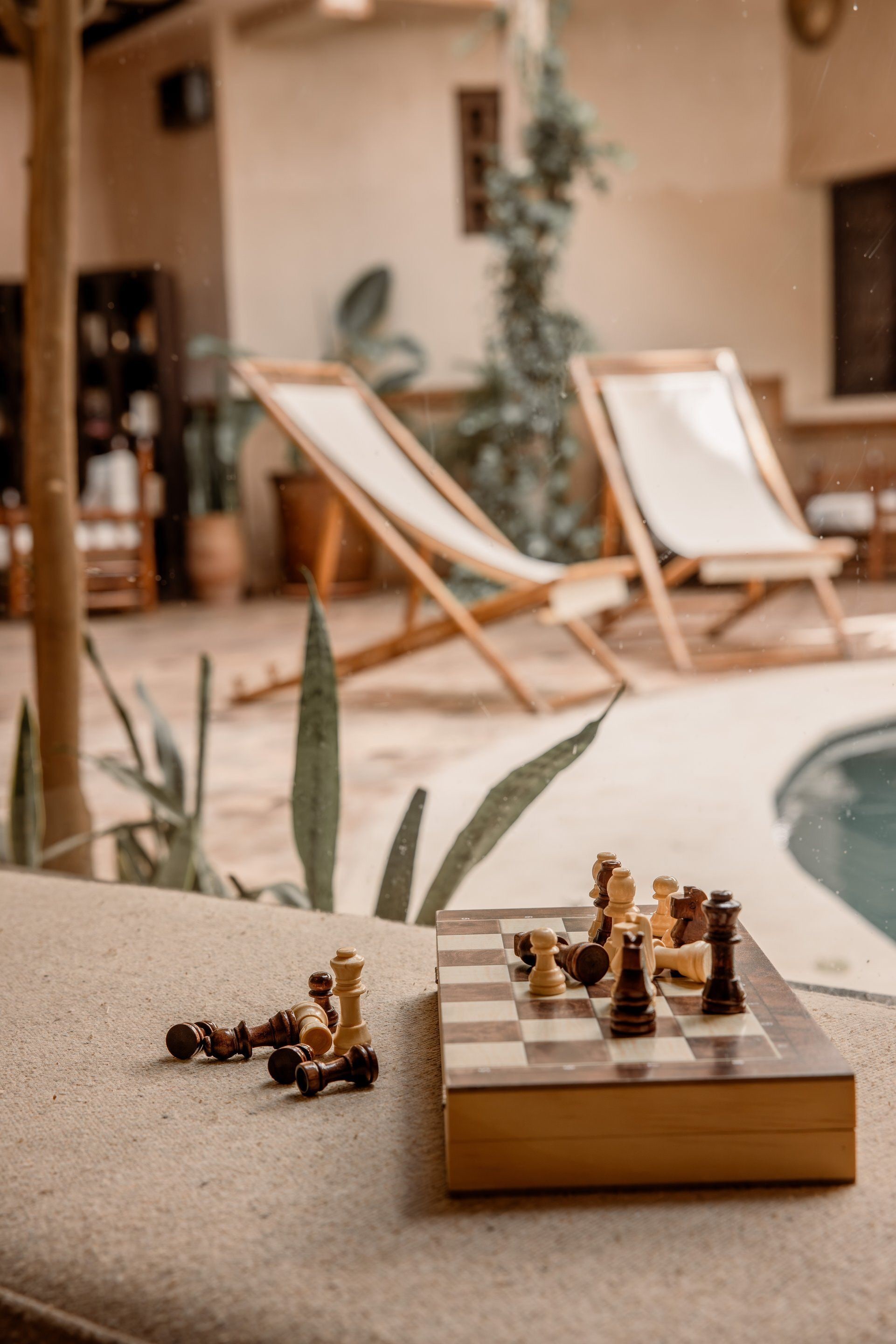 A chessboard with pieces on a textured surface, a pool, and lounge chairs in the background.