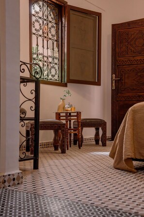 Una habitación con piso estampado, una puerta de madera con tallas elaboradas, una ventana con celosía de metal decorativa y una pequeña mesa con un florero y una vela.