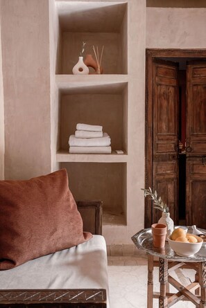A room with a sofa, a table with a bowl of fruit, and wooden shelves with folded towels. - Les Sources Berbères Riad & Spa (Marrakech)