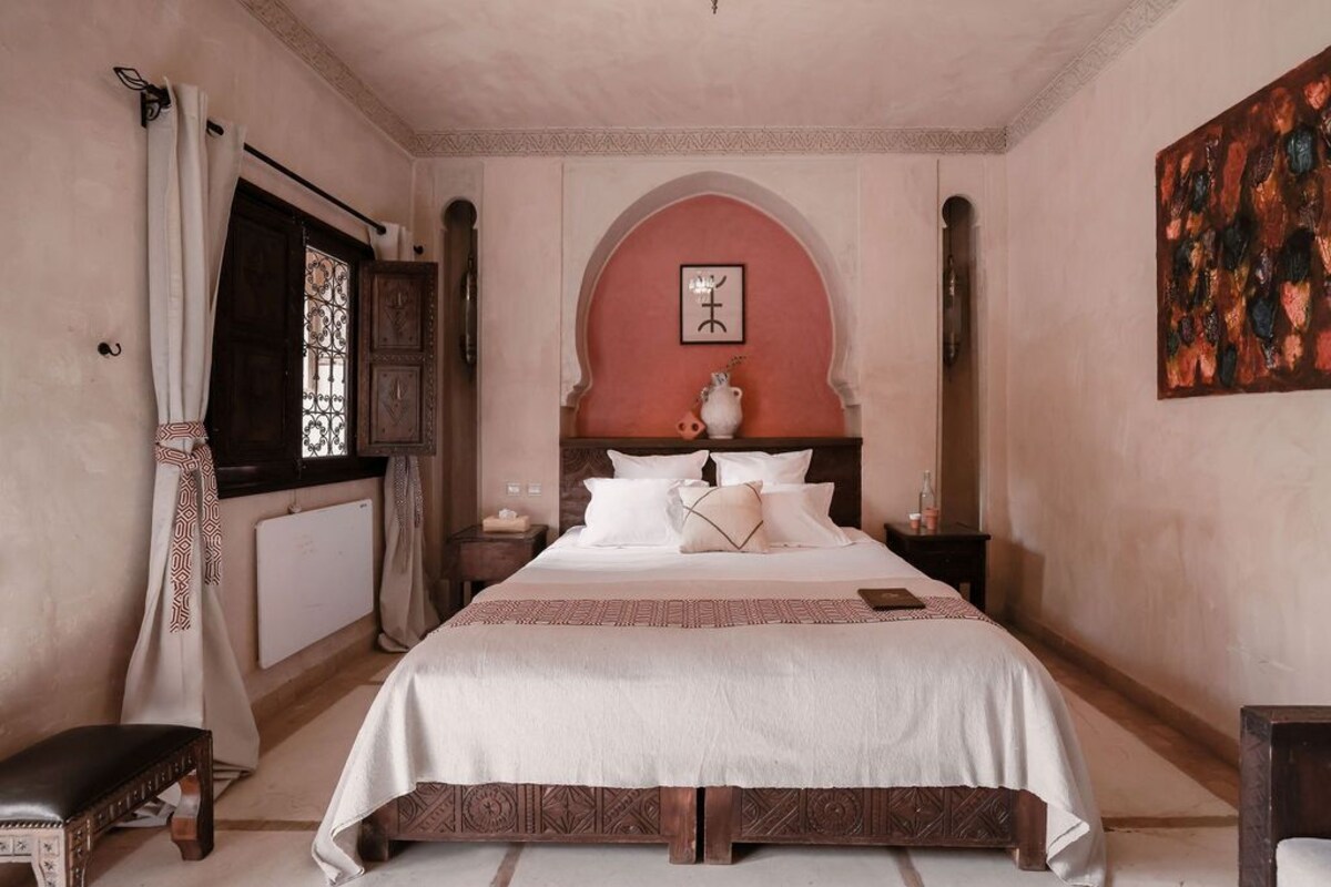 A bedroom with a large bed, a nightstand, a bench, and a window with curtains.