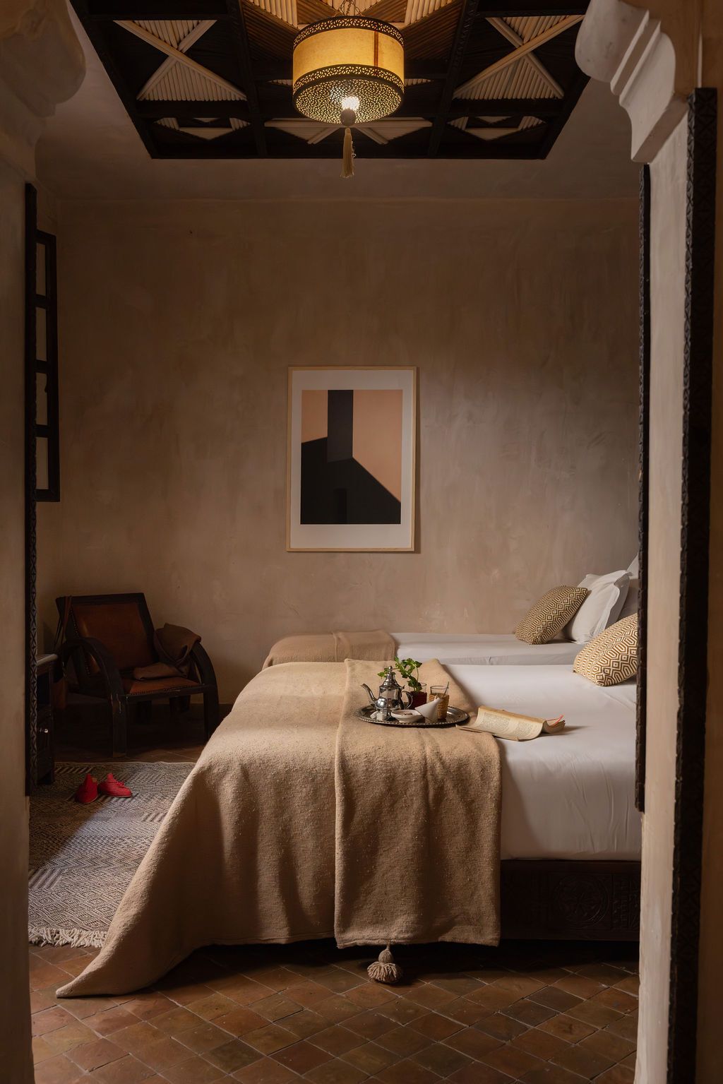 A hotel room with a bed, a chair, a table with a tray of items, and a painting on the wall.
