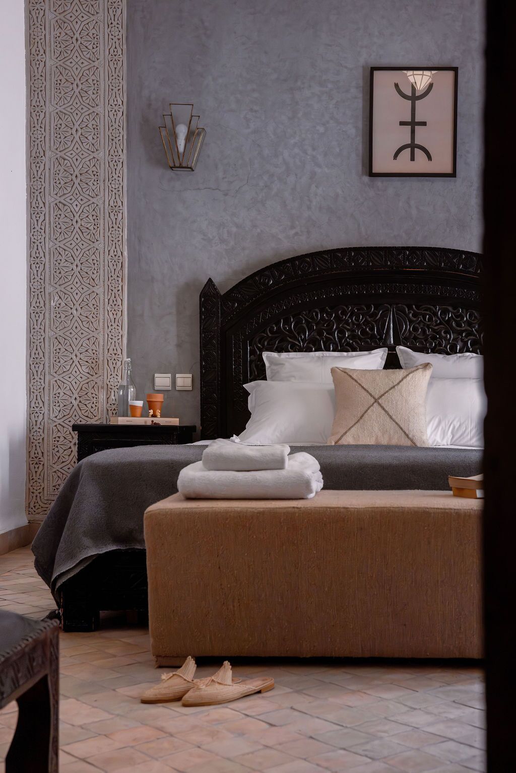 A bedroom with a dark wooden headboard, a bed with white linens, a bedside table with a lamp, and a patterned wall.