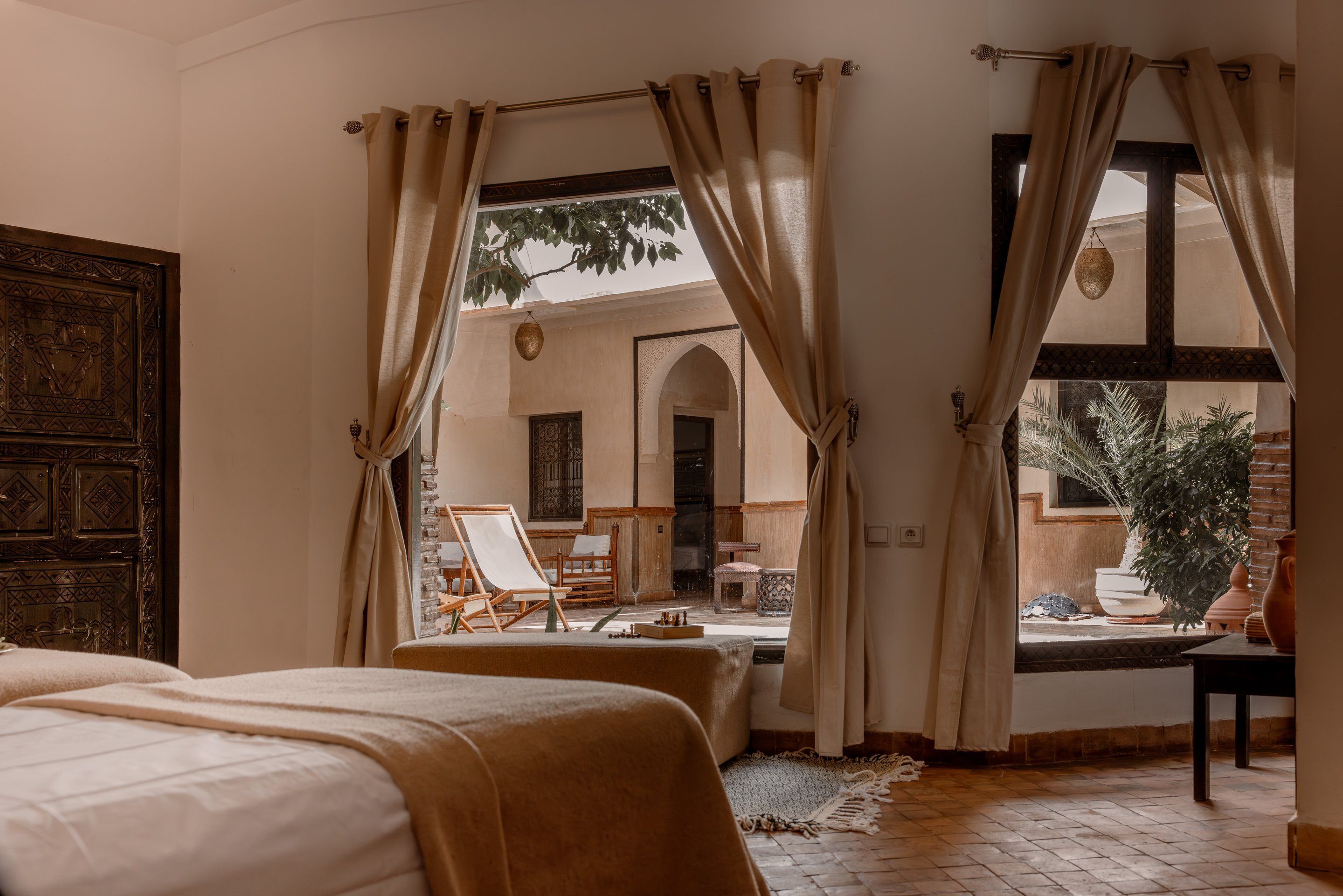 Una habitación con una cama, vista a un espacio exterior con sillas y una mesa, y una puerta de madera grande.