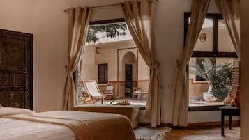 Una habitación con una cama, vista a un espacio exterior con sillas y una mesa, y una puerta de madera grande.