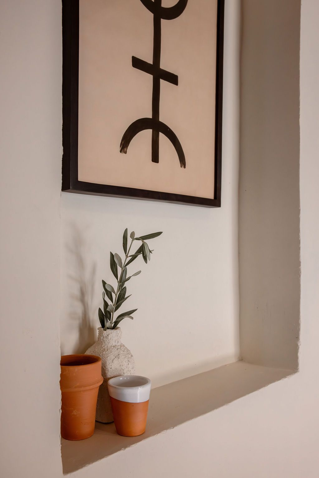 A framed artwork with a geometric design, a potted plant, and two terracotta pots on a shelf.