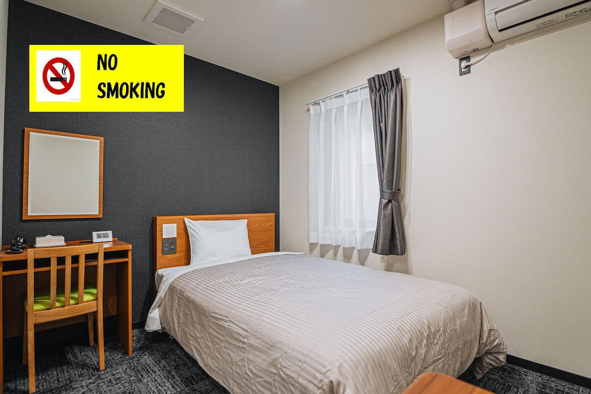 A hotel room with a bed, a desk, a chair, a "NO SMOKING" sign, and an air conditioning unit.